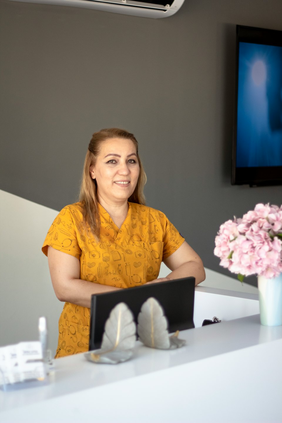 Woman in yellow scrubs smiles behind a reception desk.